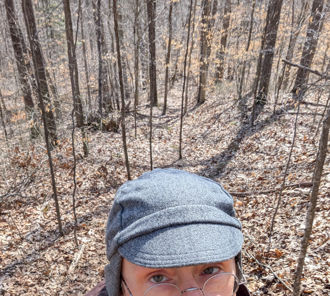 A sunny, late winter, wooded path. The bottom is the person's top part of their head, in a grey woollen cap and glasses.